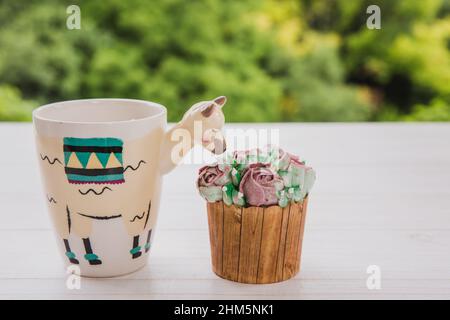 Bella tazza alla moda a forma di lama con bevanda calda e due cupcake con fiori di burro su tavolo di legno bianco con sfondo verde luminoso Foto Stock