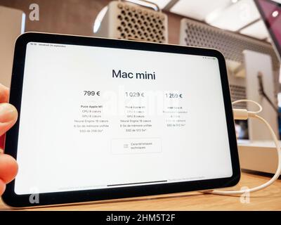 Parigi, Francia - Sep 24, 2021: POV mano maschio che tiene nuovo Mac Mini Apple Computers nuovo tablet professionale che mostra i prezzi configurazioni con mac Pro workstation computer in background Foto Stock
