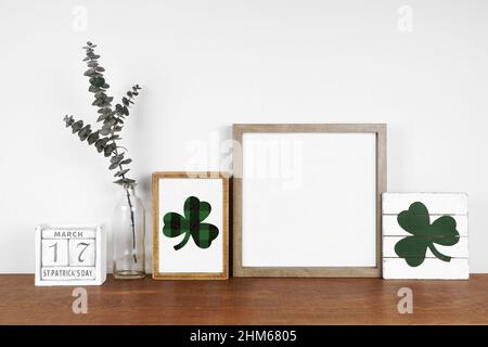 Mock up cornice in legno con St Patricks Day decor su una mensola in legno. Eleganti insegne in legno shabby, calendario e rami verdi. Cornice quadrata contro una parete bianca. Foto Stock
