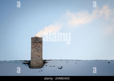Foto del tetto di una casa residenziale con un fuoco sul suo camino, utilizzato per il riscaldamento, rejeting un po 'di fumi bianchi, coperto di neve, in inverno. Foto Stock