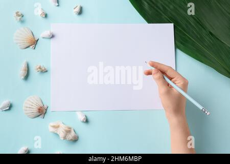 Mano femminile con matita, foglio bianco di carta e conchiglie su sfondo colorato Foto Stock