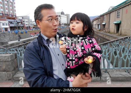 Un uomo tiene un bambino avvolto in un cappotto colorato mentre condivide un momento giocoso su un ponte su un canale panoramico fiancheggiato da storici magazzini di pietra. Foto Stock