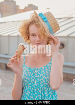 giovane allegra donna rossa ricurly allegra in cappello di paglia, zaino blu mangiando gelato sul tetto del grattacielo (tetto del tetto). Divertimento, stile di vita, urbano, moderno, Foto Stock