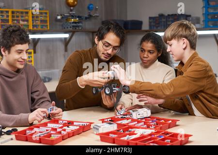 Gruppo diversificato di bambini che costruisce robot in classe di ingegneria a scuola con un giovane insegnante maschile che li aiuta Foto Stock