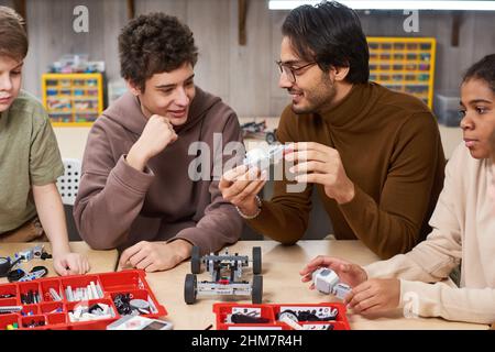 Ritratto di insegnante maschile sorridente che aiuta diversi gruppi di bambini che costruisce robot in classe di ingegneria a scuola Foto Stock