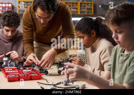 Ritratto di insegnante giovane che aiuta il gruppo diversificato di bambini che costruisce robot in classe di ingegneria alla scuola moderna, spazio copia Foto Stock