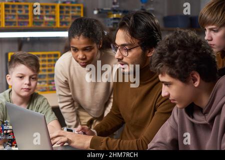 Ritratto laterale di un giovane insegnante di sesso maschile che utilizza un computer portatile con un gruppo di bambini diversi in robotica e classe di programmazione a scuola Foto Stock