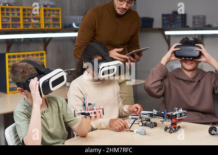 Un gruppo diversificato di bambini che indossano attrezzature per la realtà virtuale a scuola e utilizzano la realtà aumentata in classe di robotica futuristica Foto Stock