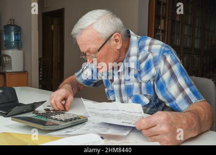 Uomo anziano seduto al tavolo e calcolando le finanze. Vecchio controllo fatture. Uomo che conta le monete sul tavolo. Concetto di calcolo della pensione Foto Stock