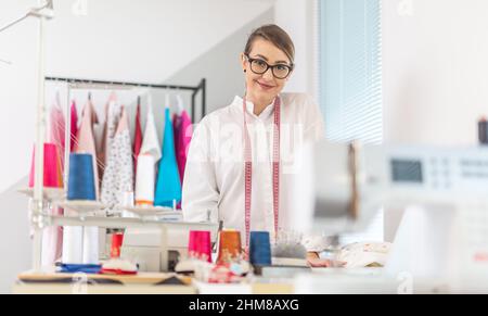 Giovane e sicura donna sarto si alza in officina con bobine di filo, macchina da cucire e appendiabiti intorno a lei. Foto Stock