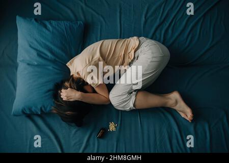 Vista dall'alto di una giovane donna a letto con pillole che giacciono accanto a lei. Una donna irriconoscibile è depressa o ha problemi mentali. Foto Stock