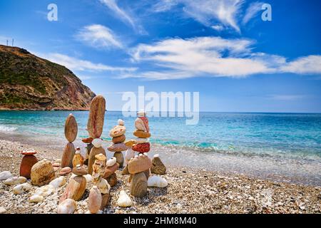 Gruppo di pietre equilibranti sulla spiaggia. Giorno di sole luminoso, atmosfera estiva, acqua di mare azzurra spruzzi su ghiaia costa, cielo blu e nuvole chiare Foto Stock