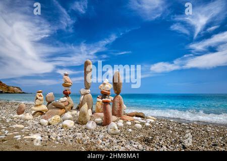 Gruppo di pietre equilibranti sulla spiaggia. Giorno di sole luminoso, atmosfera estiva, acqua di mare azzurra spruzzi su ghiaia costa, cielo blu e nuvole chiare Foto Stock