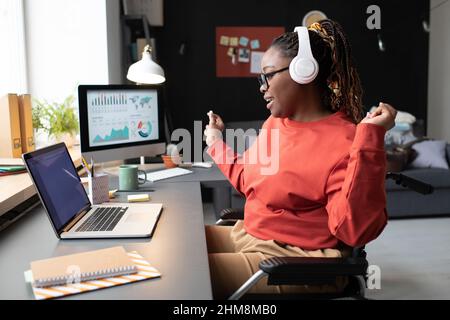 Donna africana seduta al tavolo di fronte al portatile ascoltando musica e ballando durante il suo lavoro a casa Foto Stock