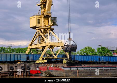 Gru fluviale portuale con benna o graniglia caricando carbone al fiume trascinare barche o chiatte ormeggiate dal molo in giorno nuvoloso. Energia, minerali, ambiente Foto Stock