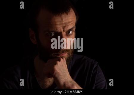 Un uomo in una maglietta scura si guarda via, appoggiando il mento sulla mano. Isolato su sfondo scuro. Foto Stock