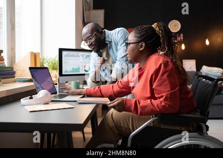 Donna con disabilità seduta su sedia a rotelle sul posto di lavoro con un laptop e discutere il progetto online con la sua collega in ufficio Foto Stock