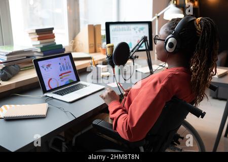 Vista posteriore della donna africana in cuffie seduta su sedia a rotelle di fronte al laptop con grafici e parlare al microfono Foto Stock