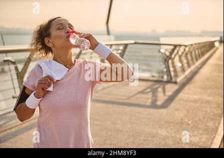 Donna sportiva di mezza età in una T-shirt rosa e leggings grigi, con braccialetti bianchi, tiene un asciugamano di spugna sulle spalle e beve acqua dopo il lavoro Foto Stock