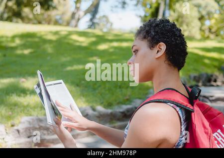 Vista del profilo di una giovane bruna forte ragazza argentina con capelli corti seduti in un parco leggendo un libro, in una bella giornata sotto l'ombra degli alberi. Foto Stock