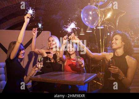 Gruppo di amici femminili che si godono festa di compleanno divertirsi con fuochi d'artificio che bevono cocktail alcolici seduti intorno al tavolo nel ristorante Foto Stock