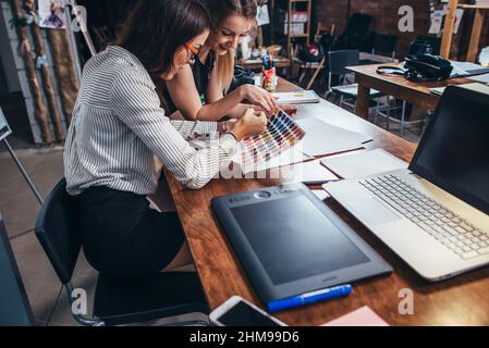 Due architetti femminili che lavorano insieme utilizzando i campioni di colore seduti alla scrivania con il laptop, tablet grafico in studio di design. Foto Stock