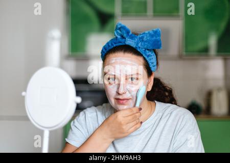 Donna massaggiante pelle del viso con maschera idratante con spazzola che indossa arco blu sulla testa guardando la fotocamera. Parlare di cura di te e della pelle. Giovani Foto Stock