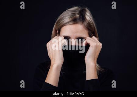 Ritratto di donna ansiosa paura che copre il volto con colletto maglione guardando la fotocamera su sfondo nero. Vittima di abuso fisico e psicologico Foto Stock