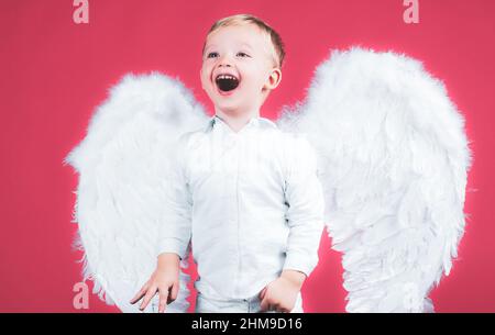 Ritratto di felice sorridente piccolo biondo riccamente Angel boy. Bambino angelico. Bambino che indossa angelo vestito bianco costume e ali di piume. Consept di Foto Stock