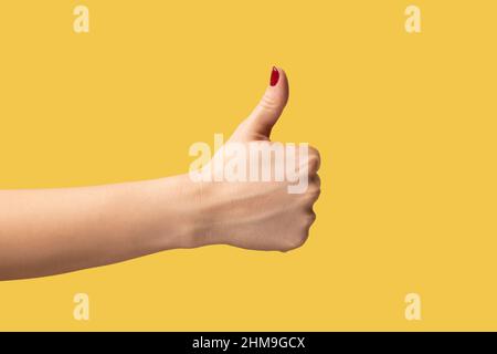 Profilo vista laterale primo piano della mano della donna che mostra come o pollici in su, approvato, feedback positivo. Studio interno girato isolato su sfondo giallo. Foto Stock