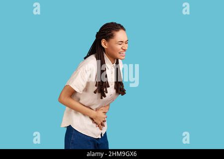 La donna dispiaciuta con i doadlock neri sente disagio nello stomaco, mantiene le palme sul pancino, ha crampi d'epoca, mangiato cibo rovinato, indossando una camicia bianca. Studio interno girato isolato su sfondo blu. Foto Stock