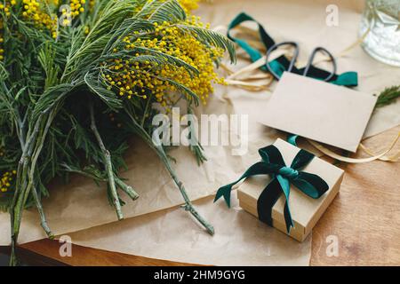 Giorno felice delle donne e giorno felice delle madri. Elegante bouquet mimosa e confezione regalo su sfondo in legno. Bella mimosa fiori in carta artigianale e regalo wit Foto Stock