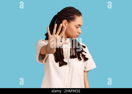 Donna gravemente infastidita con lucchetti neri che mantengono il palmo nel gesto di arresto, chiedendo di non disturbarla, guardando via, indossando una camicia bianca. Studio interno girato isolato su sfondo blu. Foto Stock