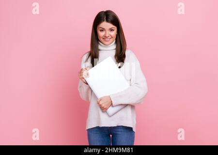 Donna bruna ottimista e positiva in piedi con notebook in mano, freelance pronto al lavoro, indossando un pullover bianco casual. Studio interno girato isolato su sfondo rosa. Foto Stock