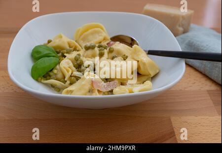 Tortellini con parmigiano, basilico, piselli e prosciutto bollito in un piatto bianco con un cucchiaio, si concentrano in primo piano Foto Stock