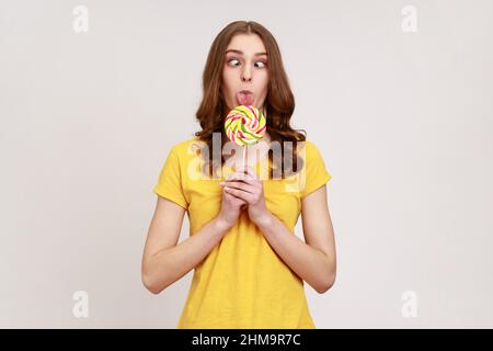 Ritratto di bambina giovane dai capelli ricci in stile urbano T-shirt leccare lecca lecca con occhi incrociati, godendo dolce, ha espressione divertente. Studio interno girato isolato su sfondo grigio. Foto Stock