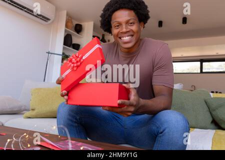 Ritratto di sorridente afroamericano giovane uomo che apre valentine dono mentre seduto sul divano a casa Foto Stock