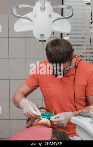 Il dentista installa una diga di gomma e un morsetto per il trattamento di un dente malato, il ragazzo è seduto in una sedia dentale. Foto Stock