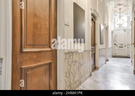 Corridoio con porte in legno e pavimento in marmo con lampadari classici luminosi appesi al soffitto ornamentale Foto Stock