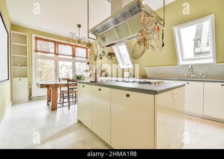 Banco con lavandino e armadietti bianchi posti vicino alla finestra in una moderna cucina luminosa con tavolo sotto lampadario luminoso in appartamento Foto Stock