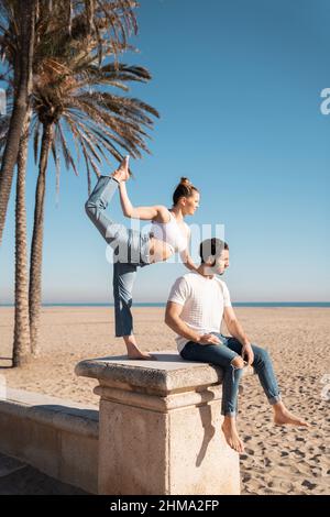 Vista laterale del corpo della donna attiva che esegue il Signore della danza asana mentre si appoggia sull'uomo seduto sulla spiaggia Foto Stock