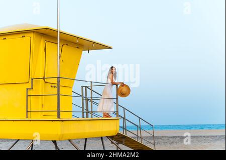 Donna viaggiatore in vestito e cappello in piedi da solo vicino cabina bagnino giallo situato sulla spiaggia di sabbia e contemplare il mare durante le vacanze estive Foto Stock