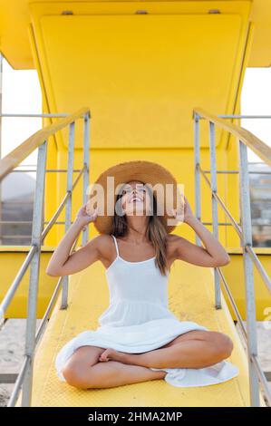 Felicissima donna in abito estivo e cappello di paglia seduto sulla torre gialla del bagnino situato sulla spiaggia sabbiosa e guardando verso l'alto Foto Stock