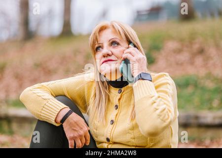 Femmina di mezza età con trucco parlando cellulare mentre si siede nel parco con alberi su sfondo sfocato Foto Stock