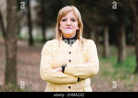 Donna di mezza età che guarda la macchina fotografica con braccia incrociate mentre si trova in piedi nel parco con alti alberi verdi su sfondo sfocato Foto Stock