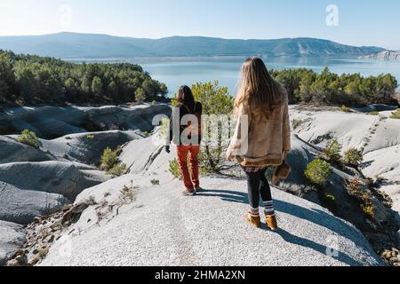 Vista posteriore del corpo di una coppia irriconoscibile in piedi su terra di pietra e ammirando il lago circondato da colline sotto il cielo blu Foto Stock