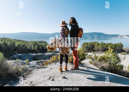 Vista posteriore del corpo di una coppia irriconoscibile in piedi su terra di pietra e ammirando il lago circondato da colline sotto il cielo blu Foto Stock