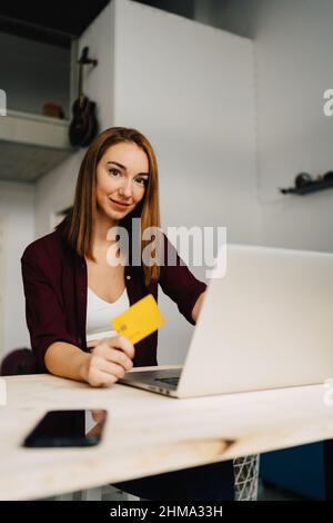 Sorridente giovane donna rossa con capelli seduti al tavolo con netbook e smartphone mentre si acquista online con carta di credito a casa e guardando la fotocamera Foto Stock