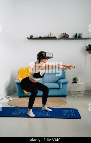 Vista laterale a tutta lunghezza di una donna sorridente irriconoscibile in abbigliamento sportivo che fa squat sul tappeto mentre fa il fitness online utilizzando il visore VR a casa Foto Stock