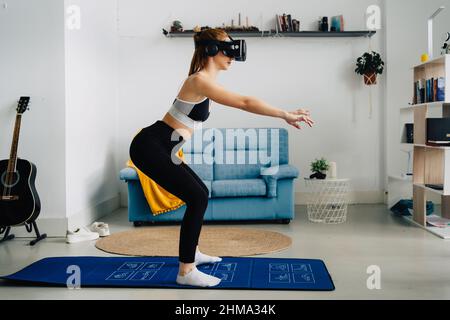 Vista laterale a tutta lunghezza di una donna sorridente irriconoscibile in abbigliamento sportivo che fa squat sul tappeto mentre fa il fitness online utilizzando il visore VR a casa Foto Stock